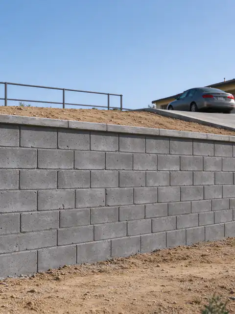 Grouted concrete block retaining wall along a sloped concrete driveway, with a metal guard rail above the drop and a car parked at the top of the drive