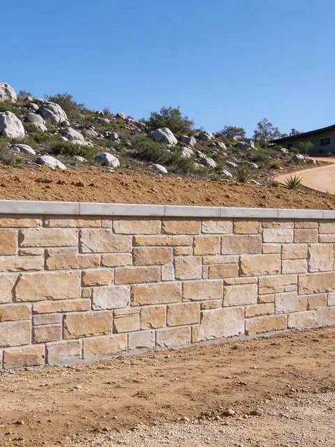 Stone-veneer retaining wall holding a cut in a boulder-strewn slope beside a driveway in Alpine
